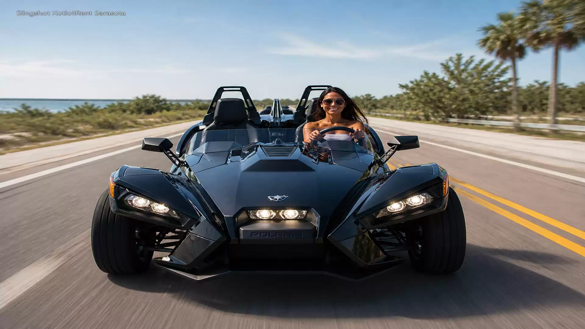 Woman driving a black three-wheeled roadster on a sunny coastal road with palm trees in the background.