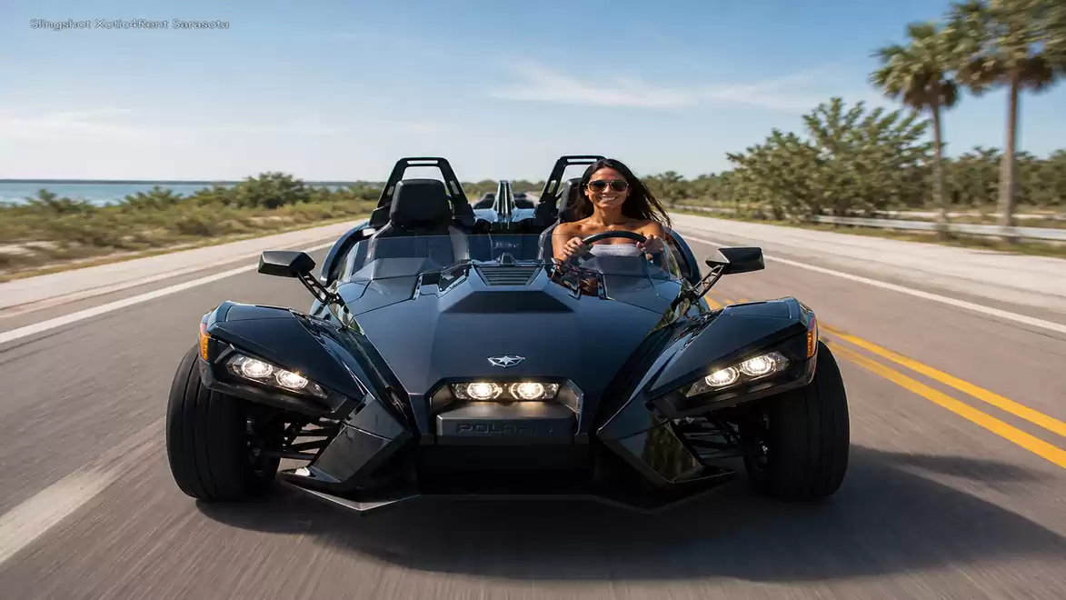Woman driving a black three-wheeled roadster on a sunny coastal road with palm trees in the background.