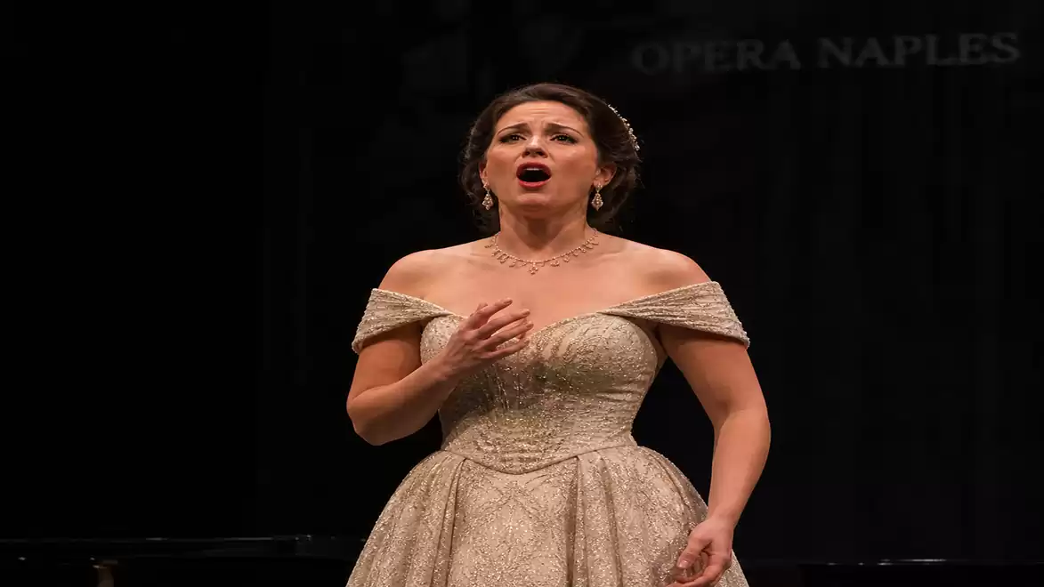 A singer in an elegant off-shoulder dress performing at an opera. The background is dark, highlighting her expressive facial expression.