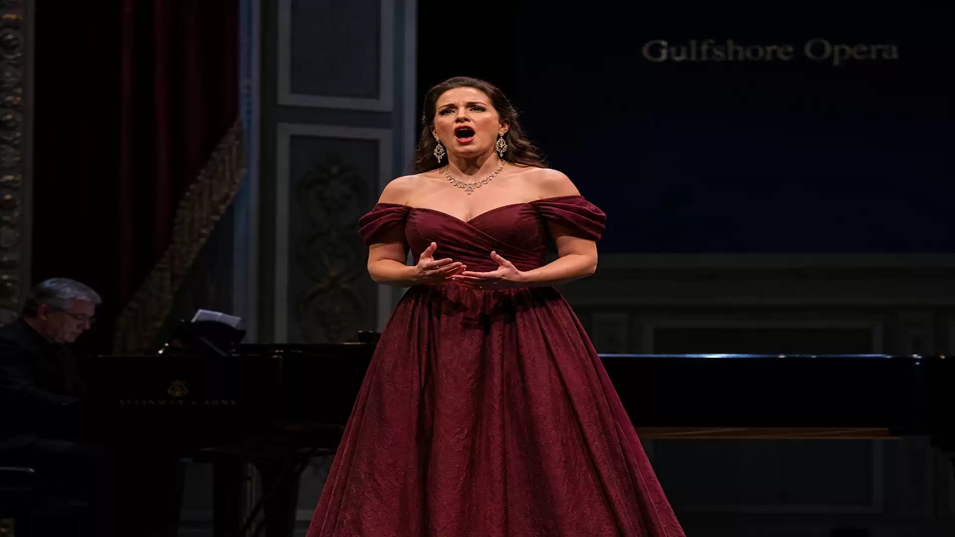 A woman in an elegant burgundy dress passionately sings on stage.
