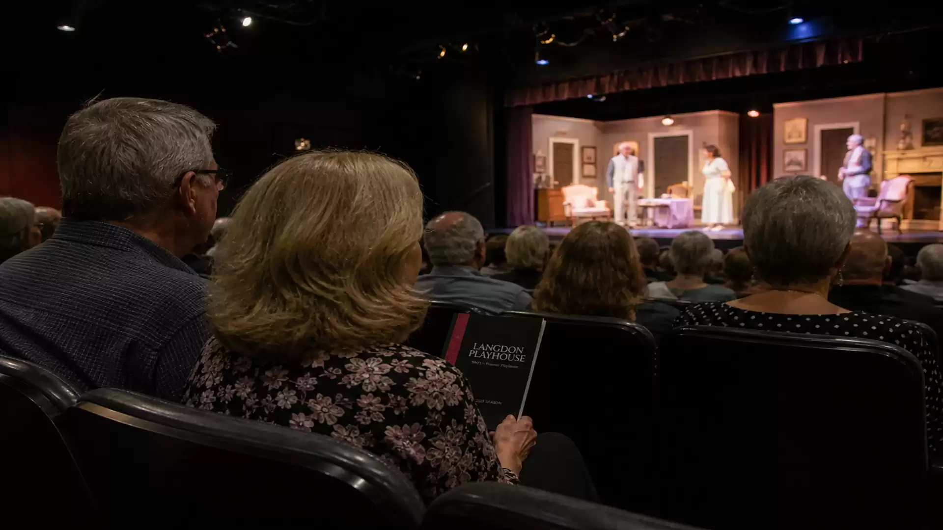 A group of people sitting in a theater watching a performance on stage. The stage is lit, and several actors are seen in action.