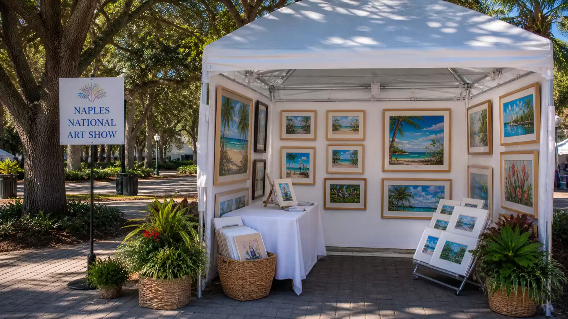 An art booth at the Naples National Art Show featuring various paintings of nature and the beach, surrounded by plants under a sunny sky.