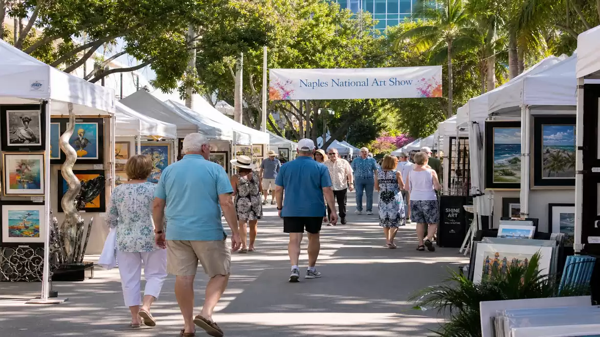 Image of an outdoor art fair with visitors walking between galleries of artwork. Bright tents and trees line the path.