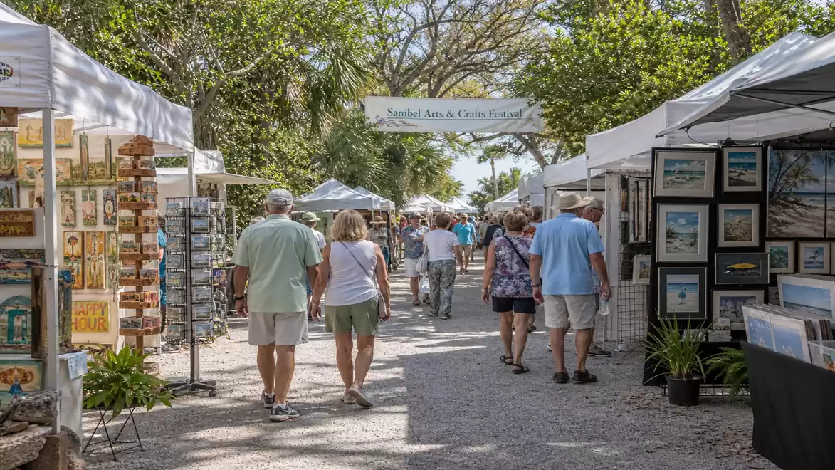 People walking through an outdoor arts and crafts market with numerous stalls, surrounded by trees.