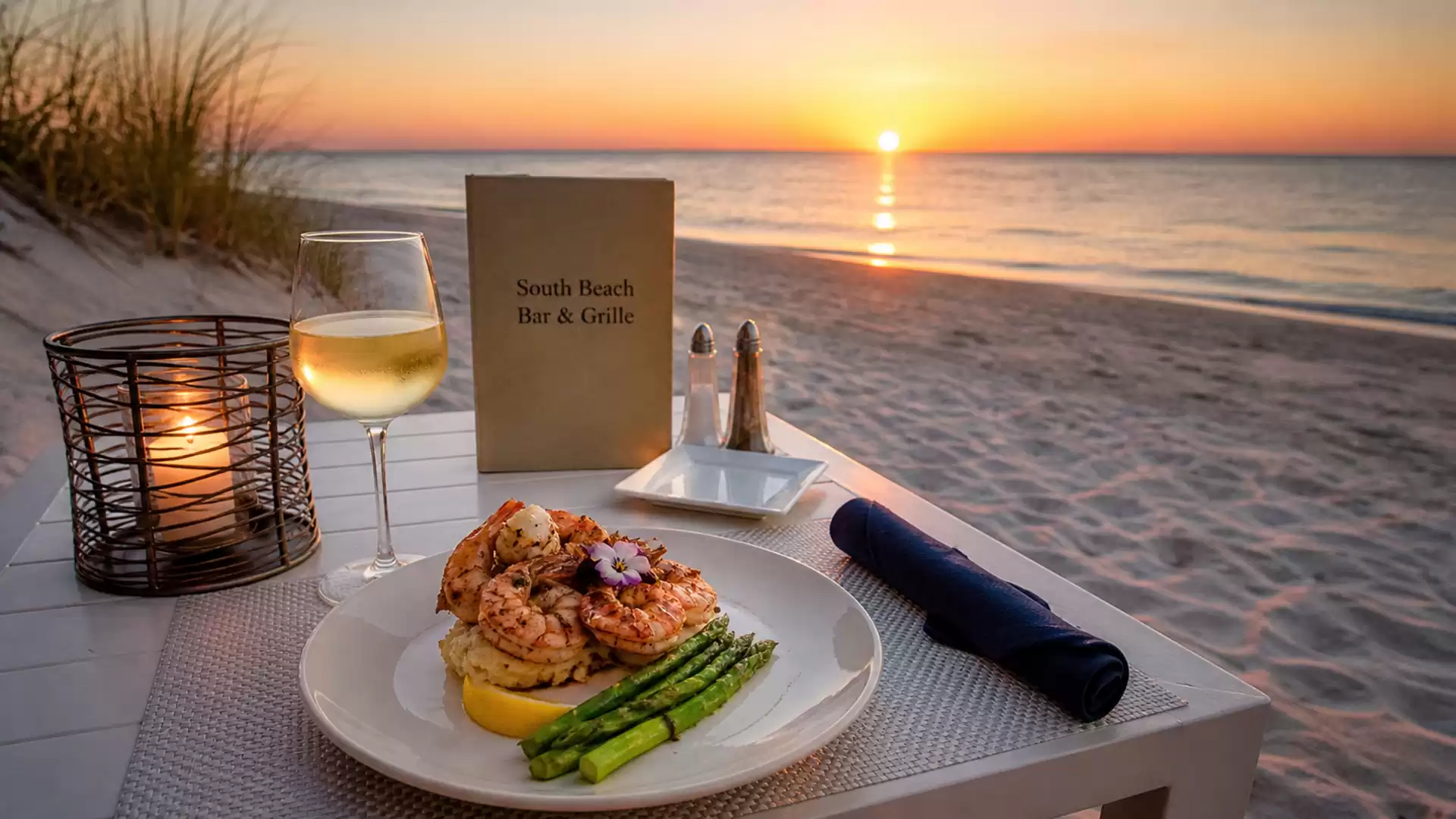 A beachside table at sunset, set with a glass of white wine and a plate of shrimp and asparagus.