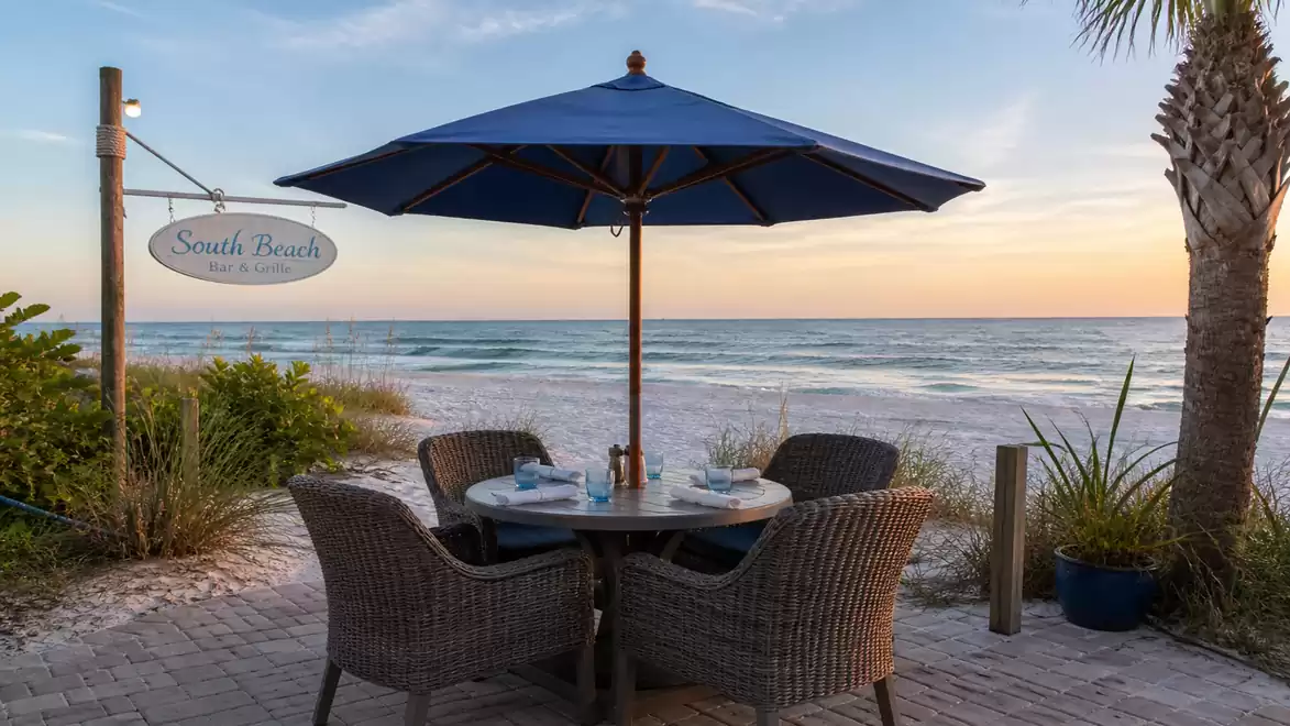 A serene beach setting with a table and chairs under a blue umbrella by the sea, surrounded by green vegetation and a sign reading 'South Beach'.