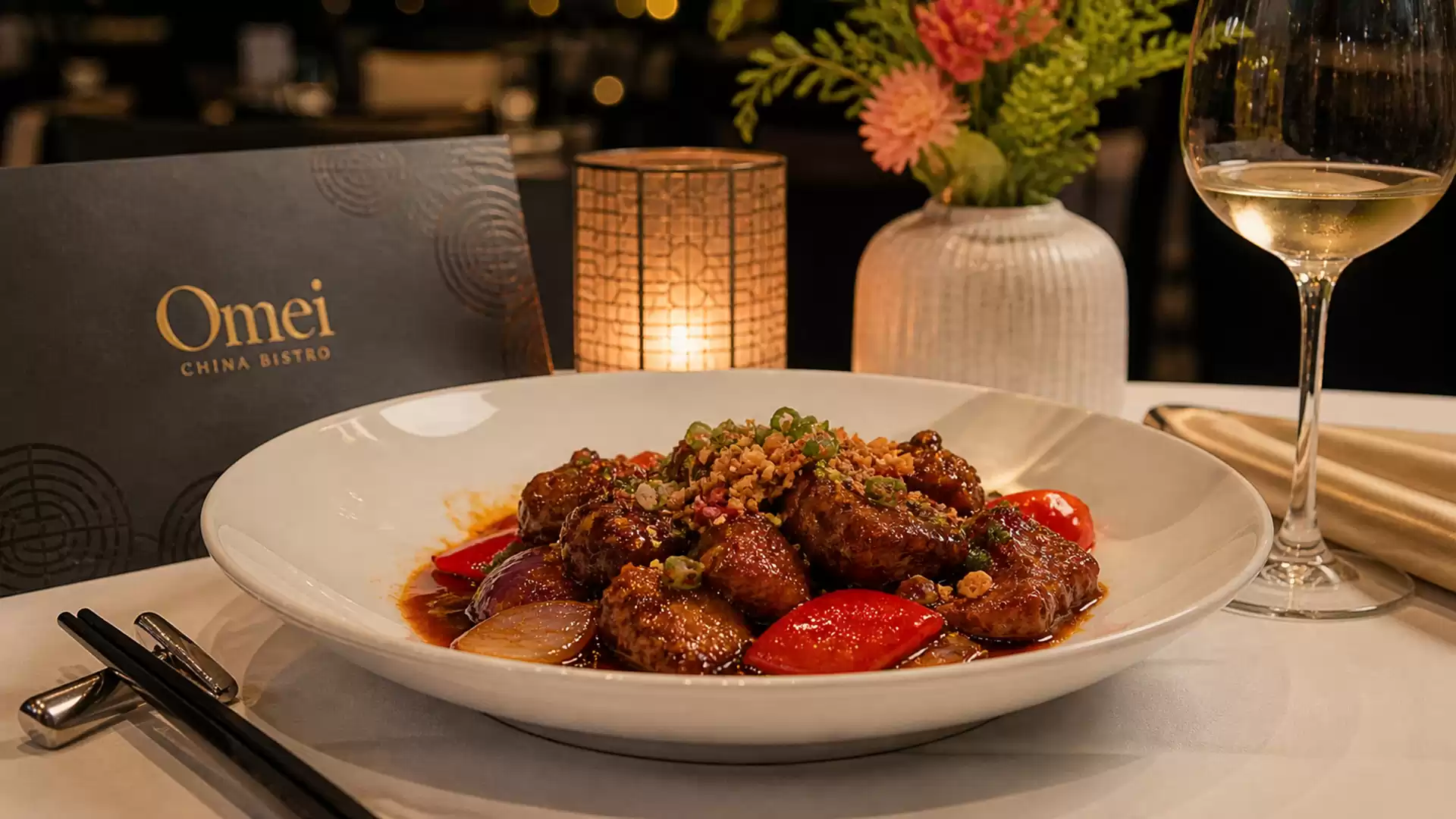 An elegant table in a restaurant featuring a plate of Asian cuisine, a glass of white wine, a menu card, and a vase with flowers.