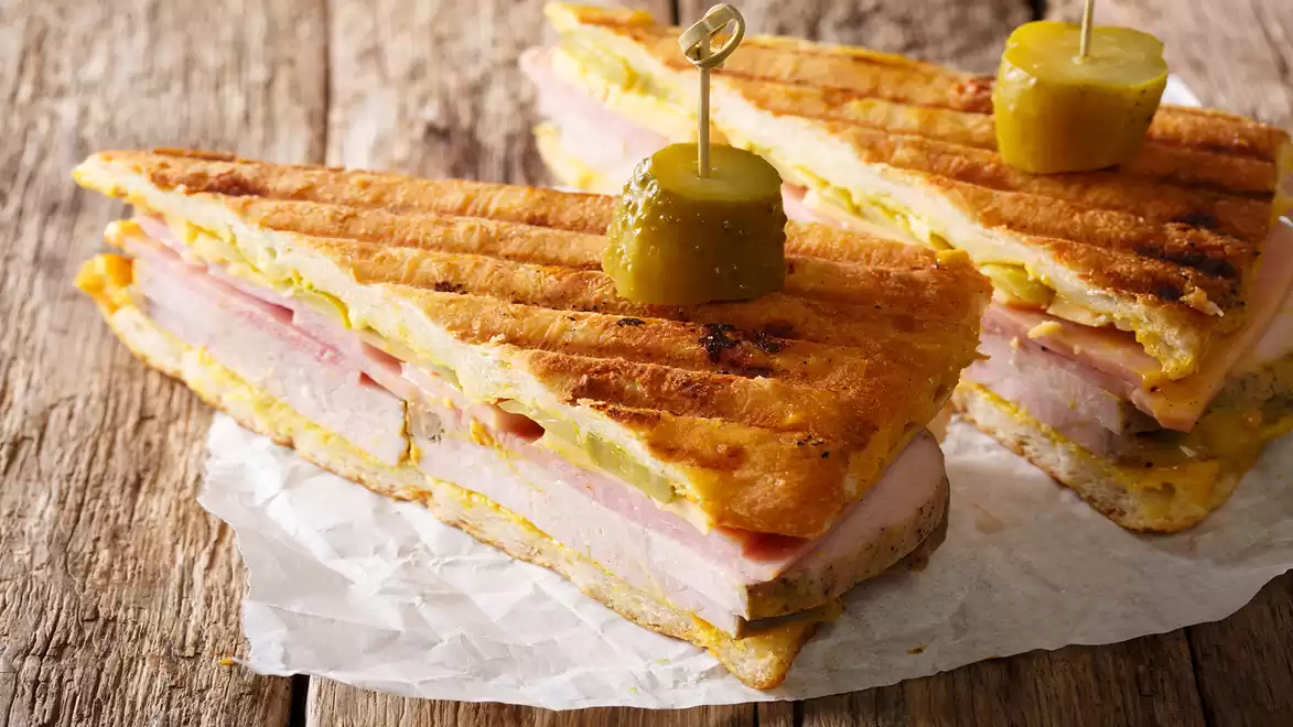 A classic Cuban sandwich with ham, cheese, and pickles on grilled bread on a rustic wooden table.