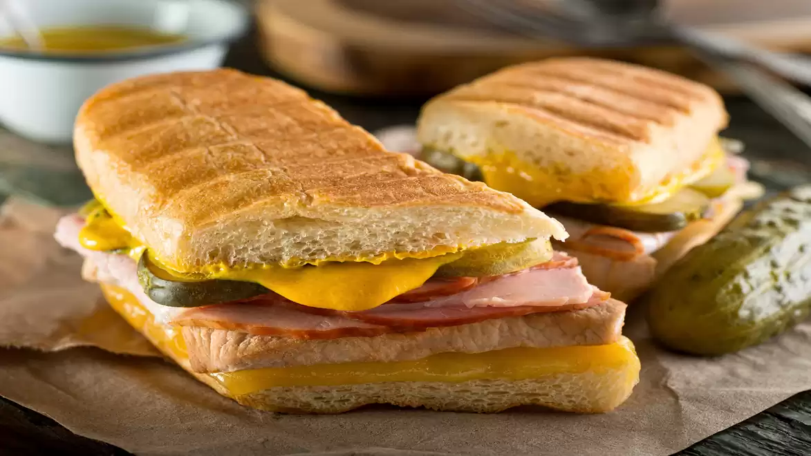 Two grilled sandwiches with ham, cheese, and pickles, served on parchment paper with a pickle in the background.