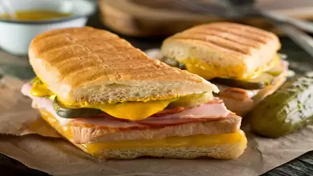 Two grilled sandwiches with ham, cheese, and pickles, served on parchment paper with a pickle in the background.