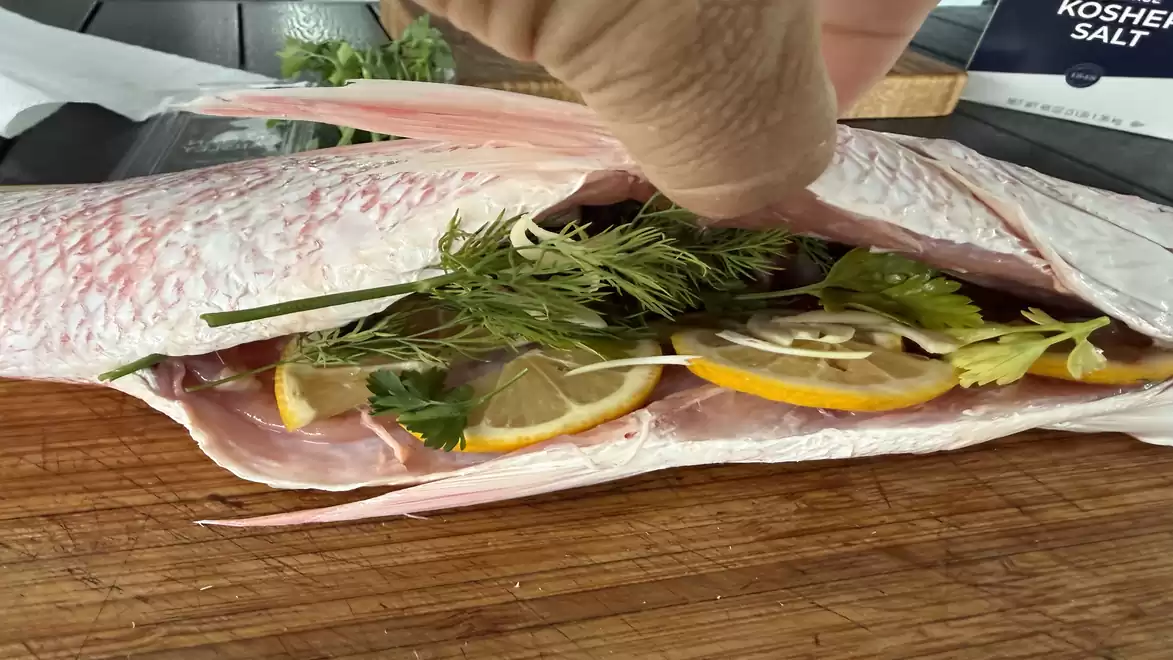 A cut-open fish fillet on a cutting board, stuffed with lemon slices and fresh herbs like dill and parsley.