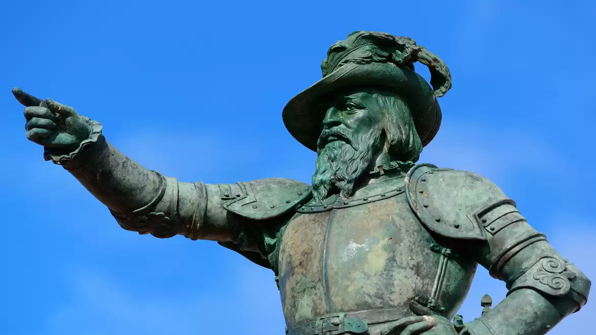 Bronze statue of a medieval knight in full armor, pointing into the distance against a clear blue sky.