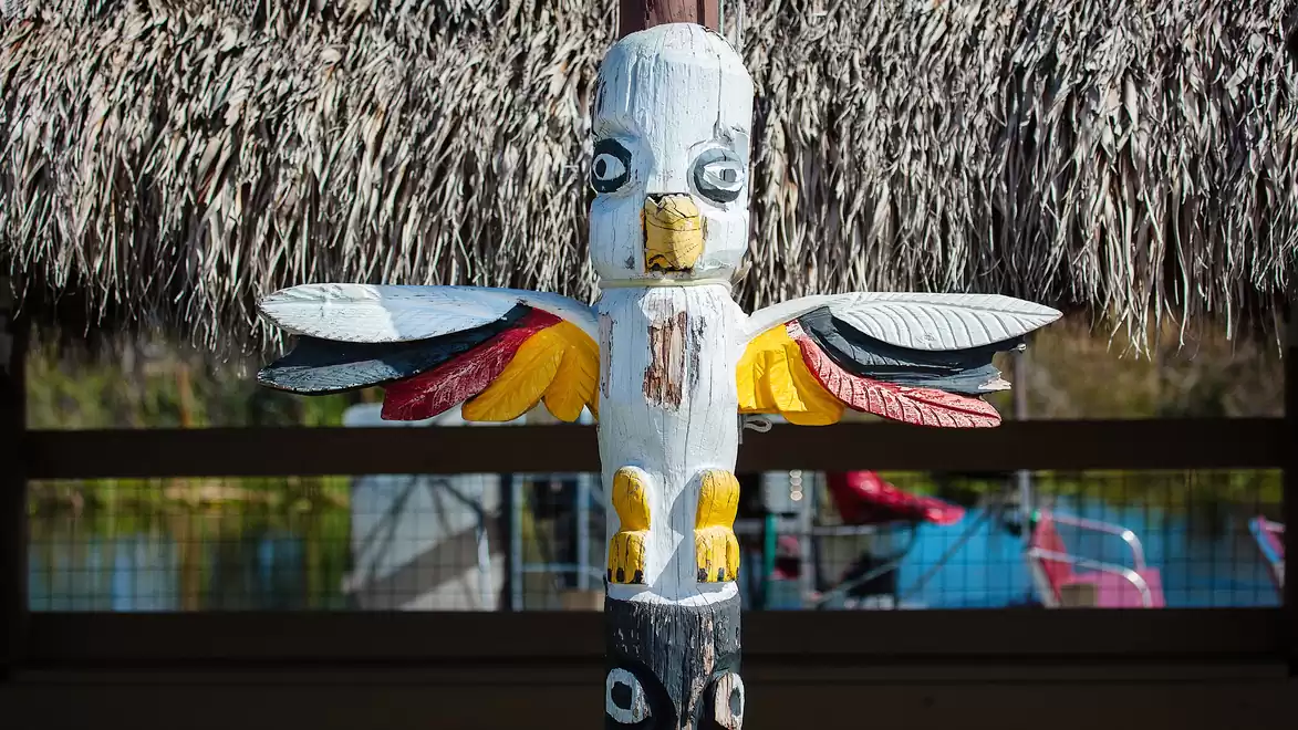 A brightly painted totem pole in the shape of a bird standing under a thatched roof.