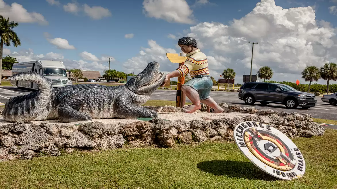 A statue depicts a person approaching an alligator. The scene is set on a grassy area next to a road under clear skies.
