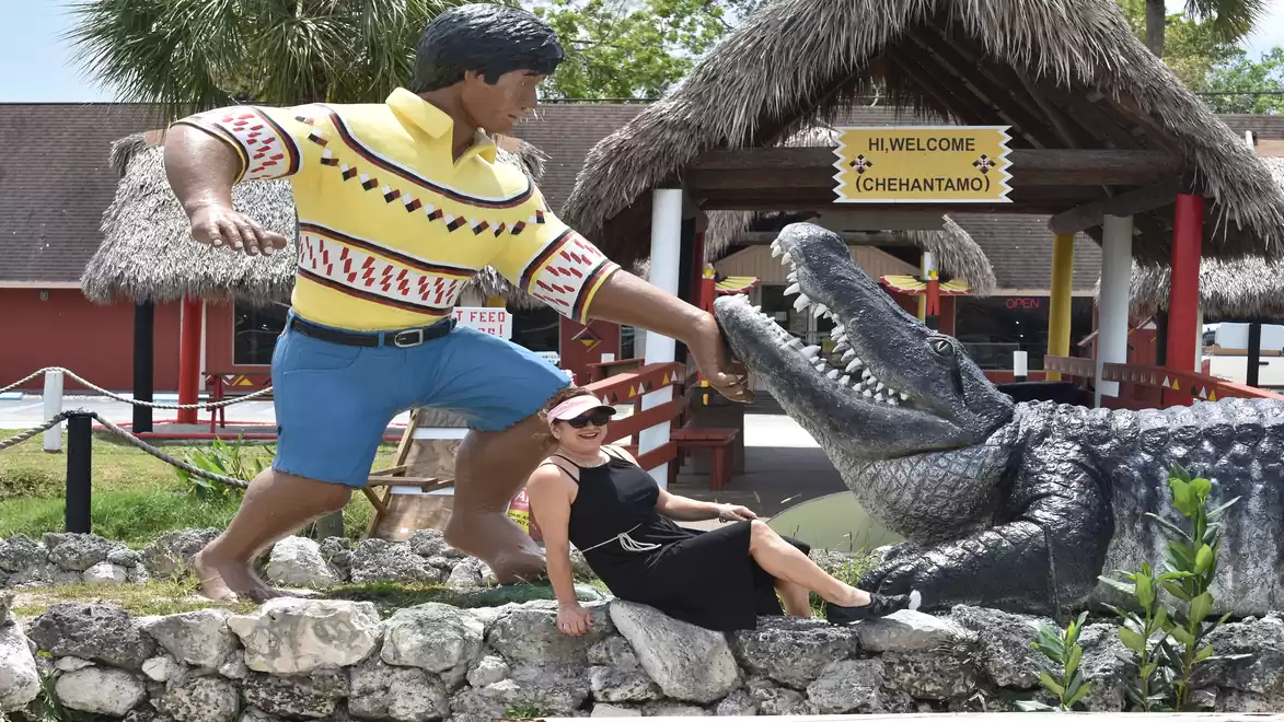 A woman sits in front of a large alligator statue and a statue of a man in shorts and a shirt at a tourist attraction with a sign 'Welcome Chéhantamo'.