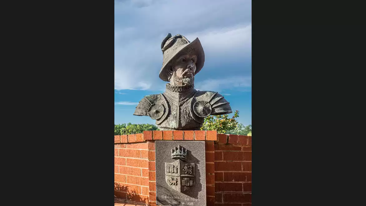 Bronze statue of a man in Renaissance attire with a hat, mounted on a brick pedestal with a coat of arms, set against a blue sky.