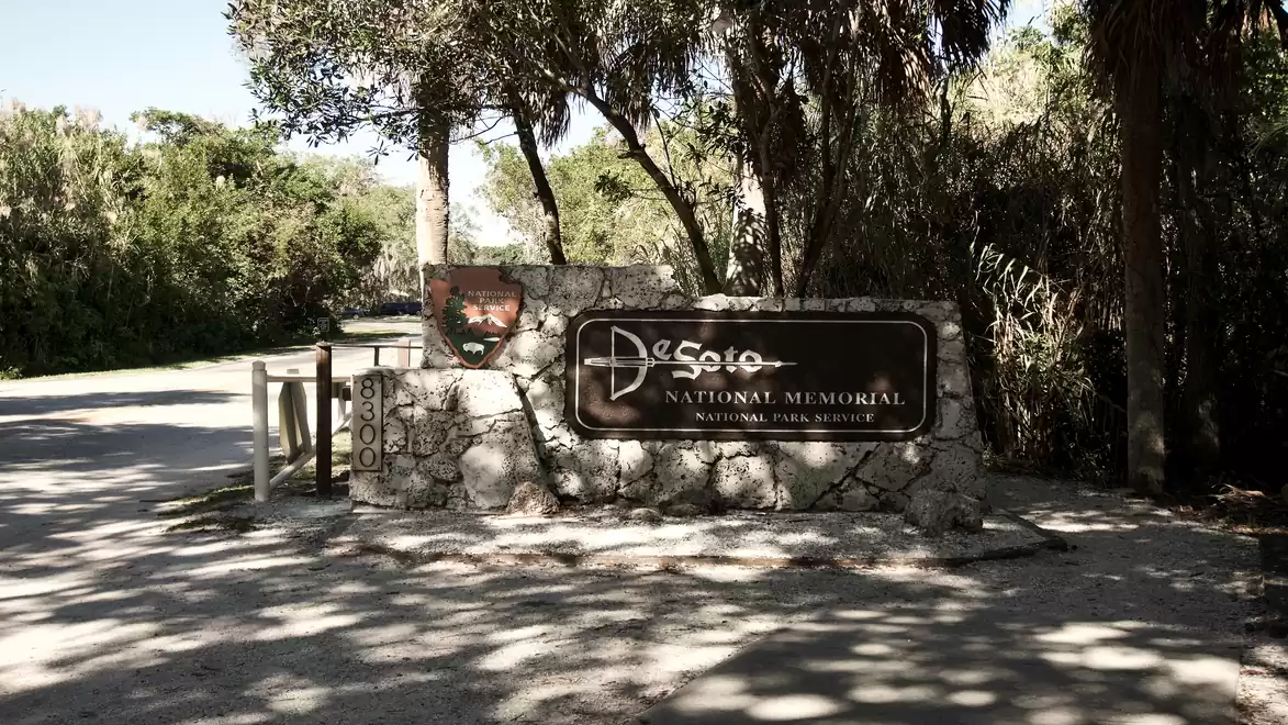Entrance sign of DeSoto National Memorial surrounded by trees and shadows.