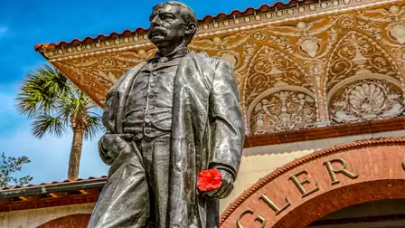 Statue of Henry Flagler in front of an ornate building with palm trees in the background, holding a red rose.