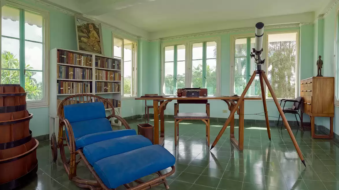 A bright room with a telescope beside a wooden desk and a bookshelf in the background.