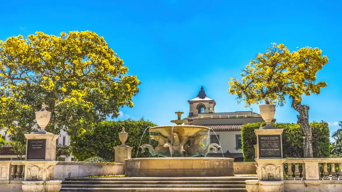 Fountain in a sunny park with trees and a historic building in the background.