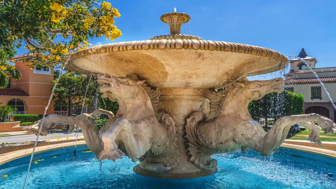 An impressive outdoor fountain with decorative stone horse sculptures protruding from the round basin, surrounded by lush greenery and a bright blue sky.