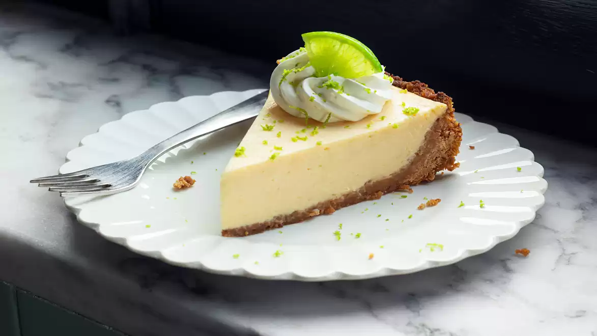 A slice of lime cheesecake with whipped cream topping and lime decoration on a white plate with a fork on marble surface.