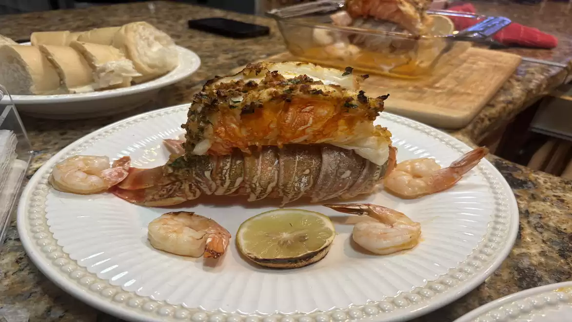A beautifully presented plate with grilled lobster and shrimp, garnished with a lemon slice. In the background, more food is visible on a countertop.