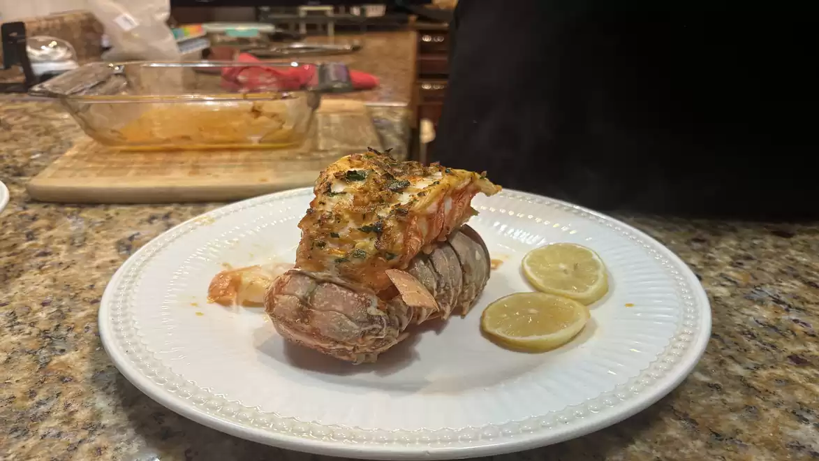 Grilled lobster on a white plate with lemon slices, kitchen utensils in the background.