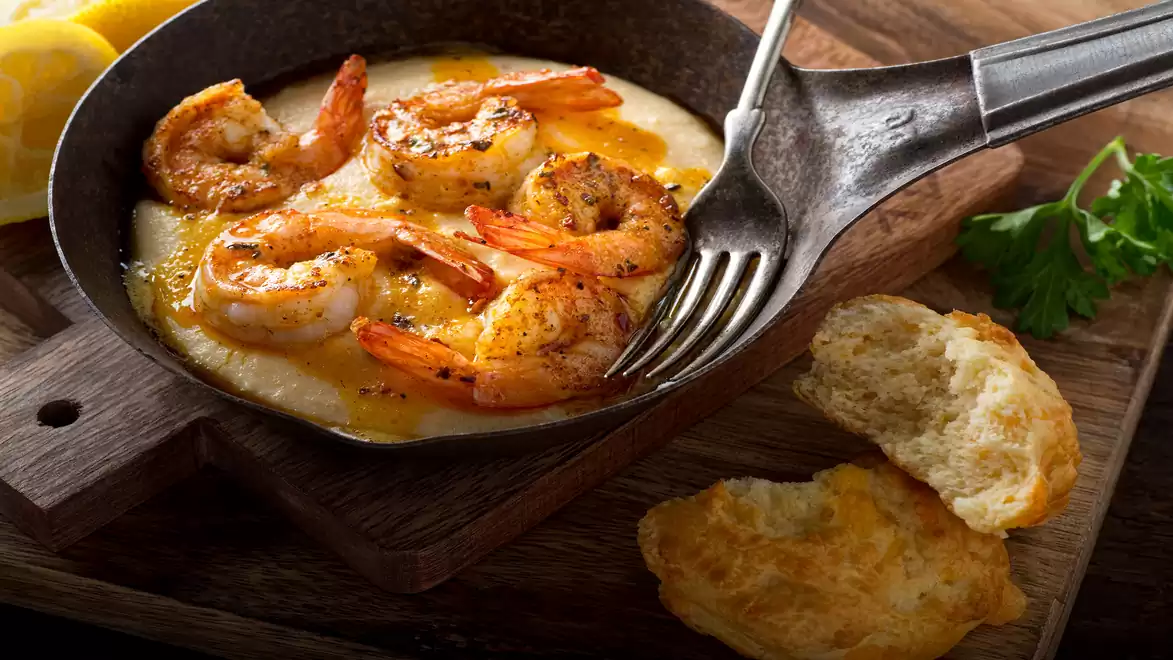 A plate of creamy polenta topped with grilled shrimp, served in a cast iron skillet. Fresh herbs and bread are placed alongside on a rustic wooden board.