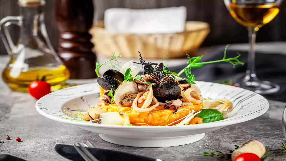 An elegant gourmet dish with black pasta, seafood, and fresh herbs, served on a white plate, arranged on a rustic table with a wine glass in the background.