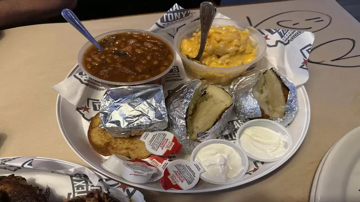 A plate with beans, mac and cheese, a baked potato in foil, toast, and sour cream. A typical American BBQ dish.
