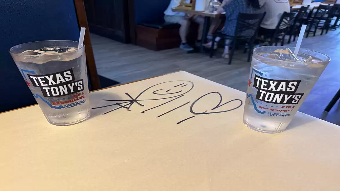 Two drinking glasses with Texas Tony's logo on a table, with a drawing in the background.