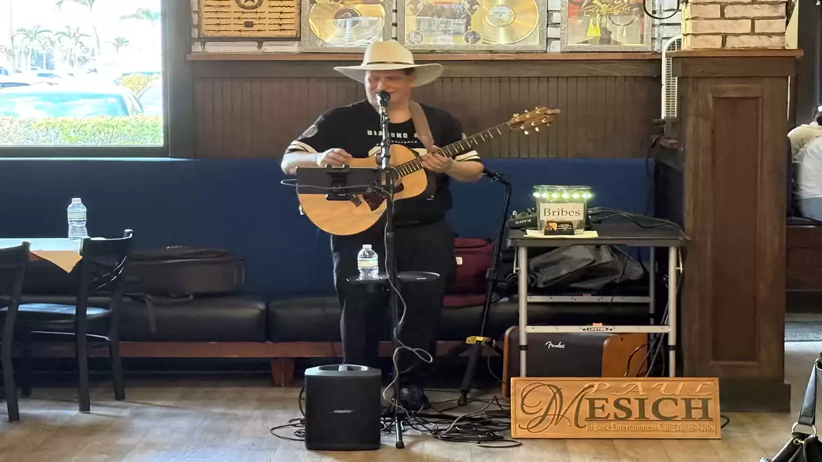 A musician wearing a cowboy hat plays guitar in a restaurant, standing behind a microphone, surrounded by music equipment.