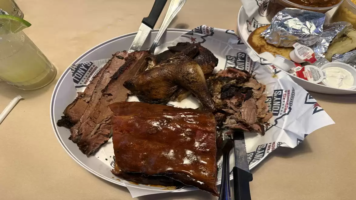 A plate with various grilled meats, including beef, chicken, and pork ribs, arranged on printed paper. Sides like grilled bread and sauces are visible next to it.
