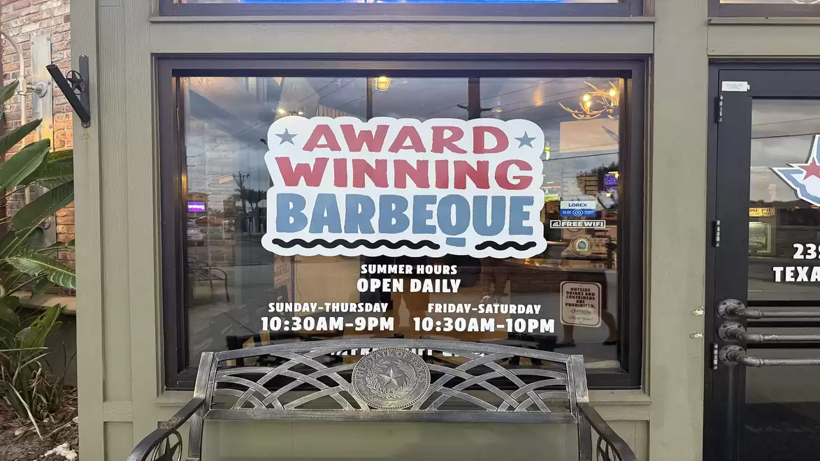 Entrance of a restaurant with a large sign saying 'Award Winning Barbecue'. Summer hours are Sunday-Thursday from 10:30 AM to 9 PM and Friday-Saturday from 10:30 AM to 10 PM.