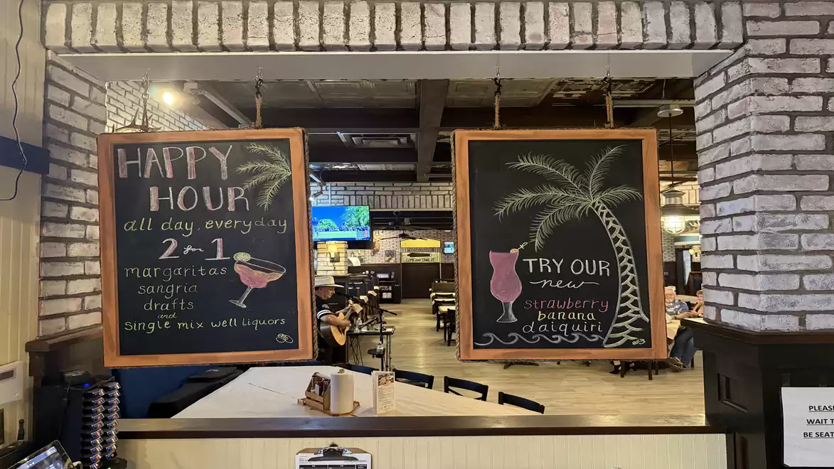A restaurant bar with two chalkboards advertising special drinks and happy hour offers. The left board features deals for Margaritas and Sangria, while the right board showcases a strawberry banana daiquiri under a drawn palm tree.