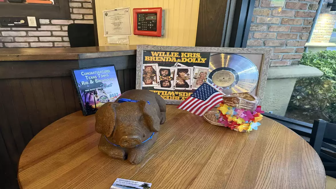 A round wooden table with a gold record, a dog statue, a small US flag, and decorative items in a room with a brick wall.