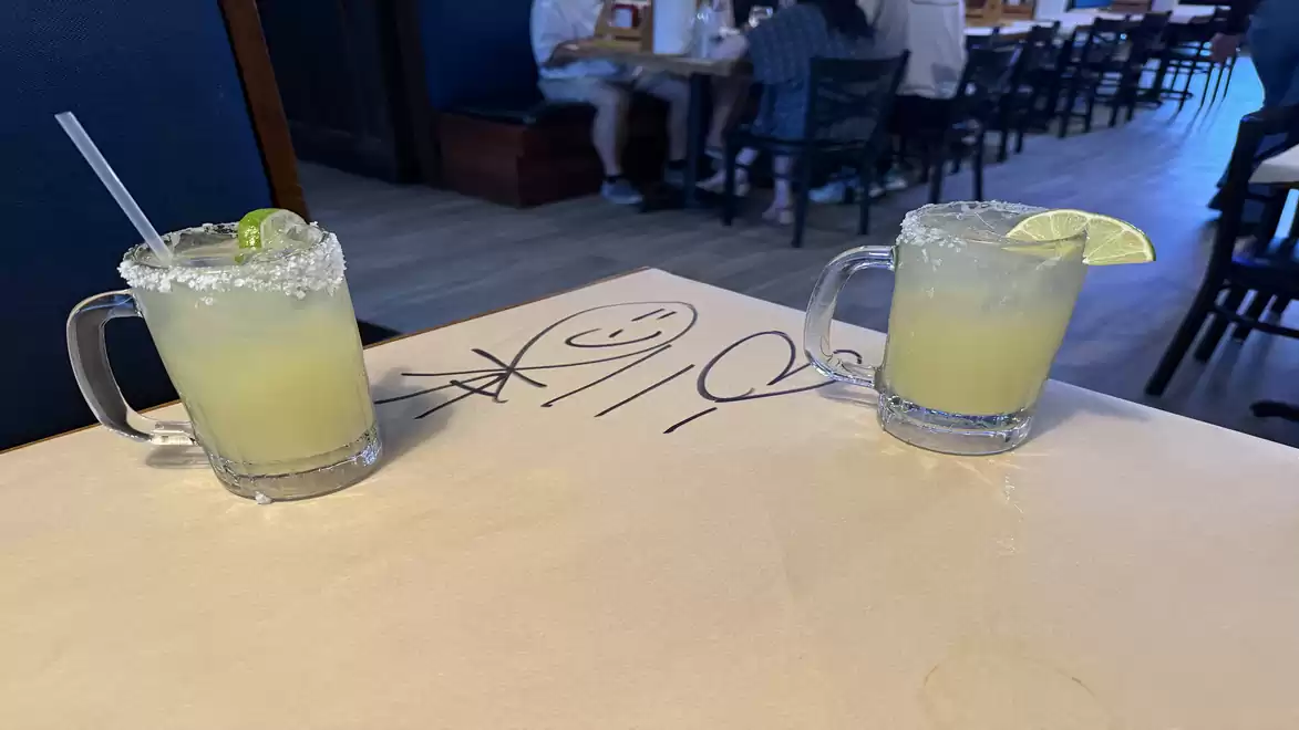 Two glasses of margarita with salted rims and lime wedges are on a table in a restaurant. Blurry people are visible in the background.