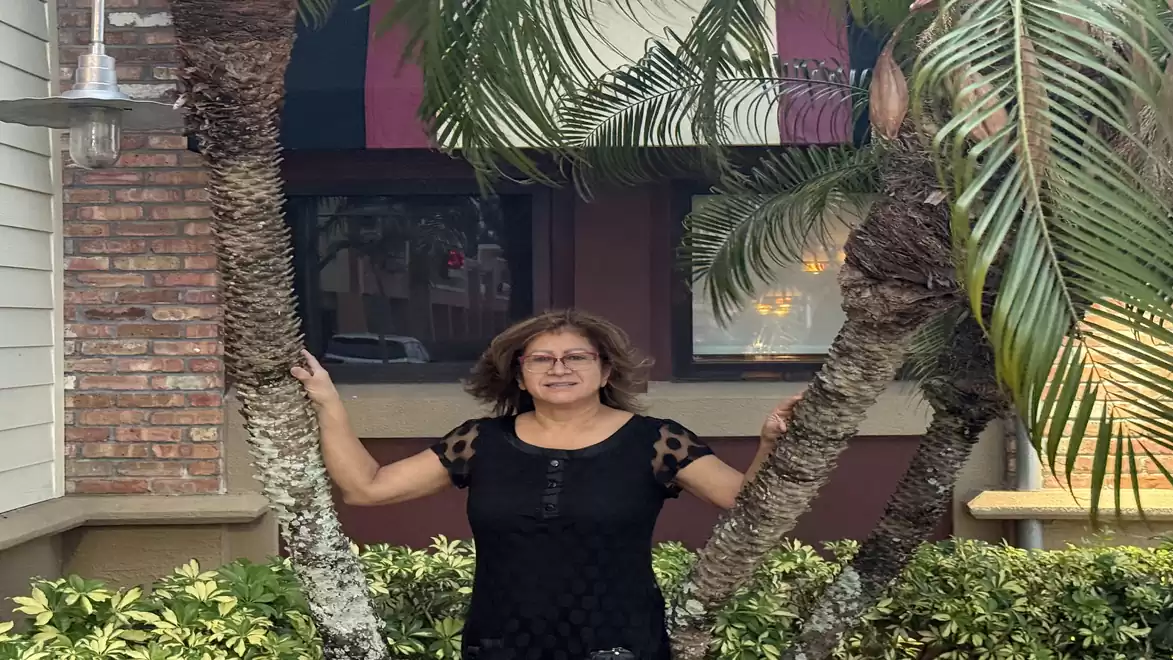 Woman stands between two palm trees in front of a brick house.
