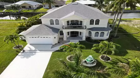 Aerial view of a large white house with a manicured garden and fountain, surrounded by palm trees in a residential area.