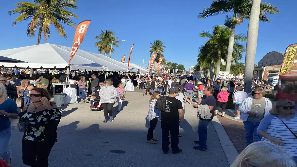 Open-air market on a sunny day with many people, palm trees, and white tents.