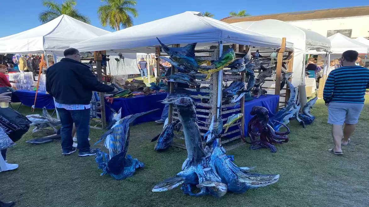 Outdoor art market with tents and decorative brightly painted wooden sculptures under a blue sky.
