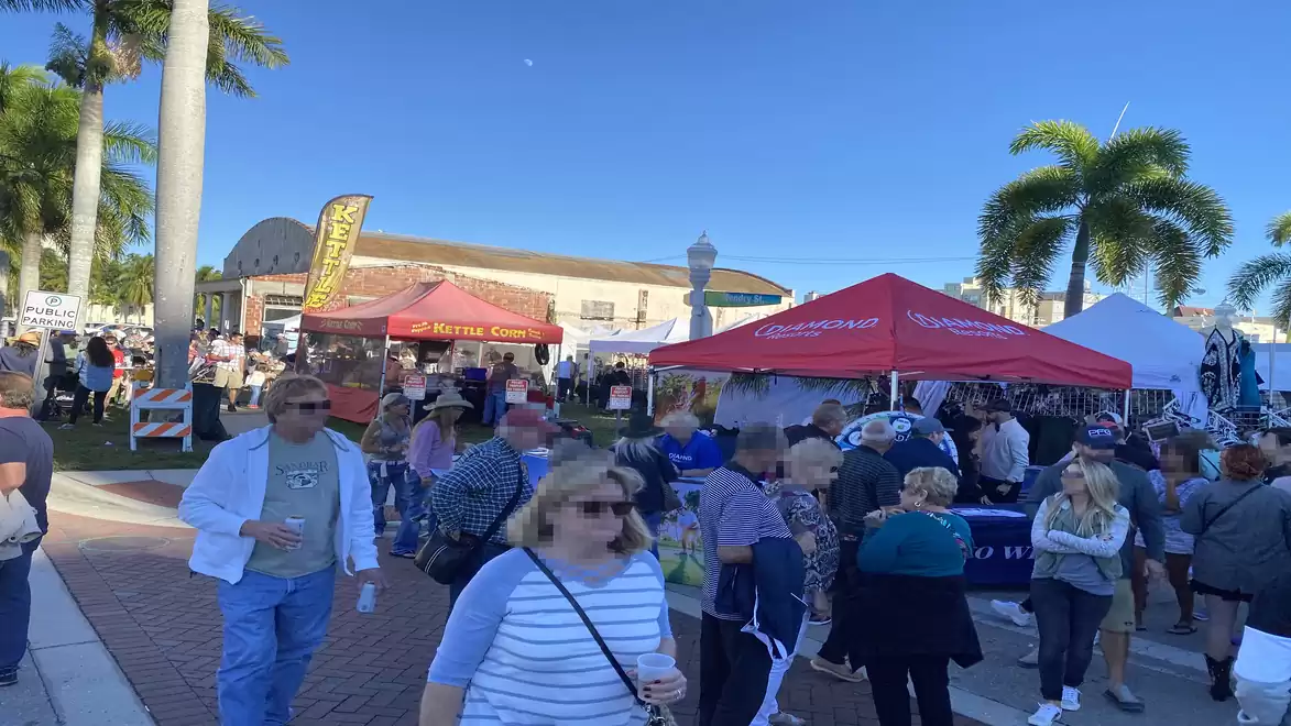 An outdoor festival with many people walking around various food stalls under red and white tents. Palm trees in the background and a clear blue sky emphasize the sunny weather.