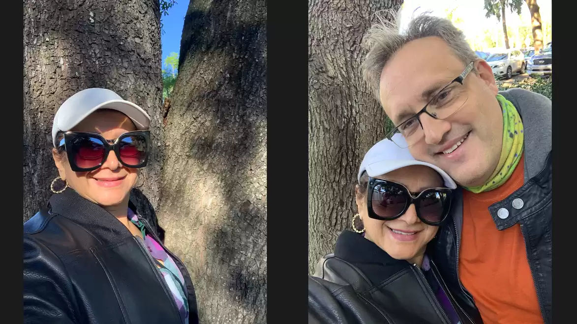 A woman wearing sunglasses and a cap takes a selfie in front of a tree. Next to her is a man with glasses and a scarf. Both are smiling at the camera.