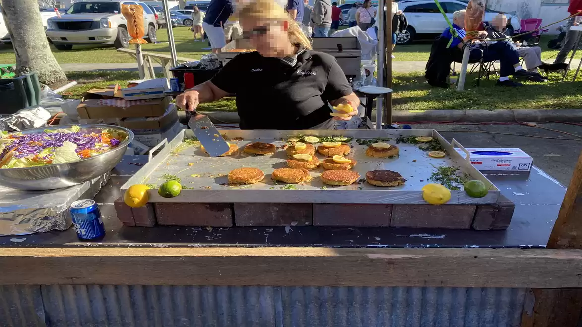 A person grilling burgers and vegetables on a large outdoor griddle at a street festival. Limes and lemons are visible in the foreground.