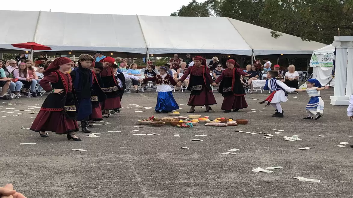 Group of children in traditional costumes dancing at an outdoor event while spectators watch under a tent.