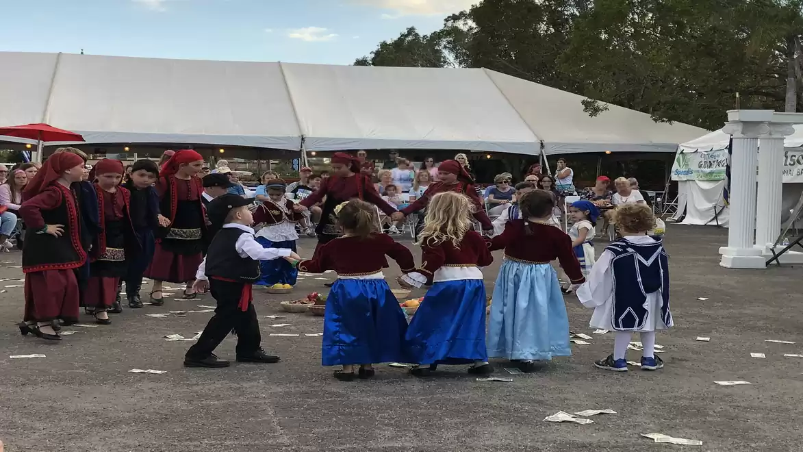 Children in traditional costumes dancing in a circle at a festival with a large tent in the background.