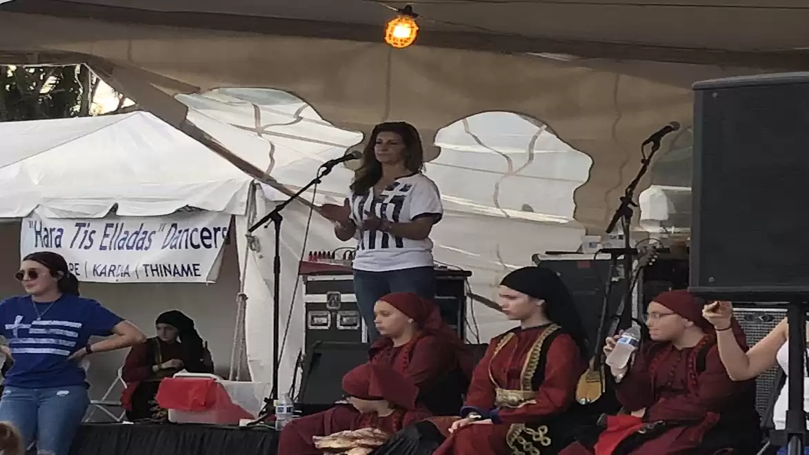 A group of people on a festival stage. In the front, young dancers in traditional clothing are seated, while a woman speaks into a microphone in the background.