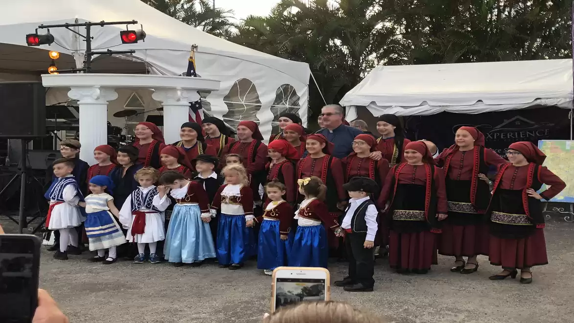 A group of children and adults in traditional clothing smilingly posing in front of a festival tent, surrounded by trees.