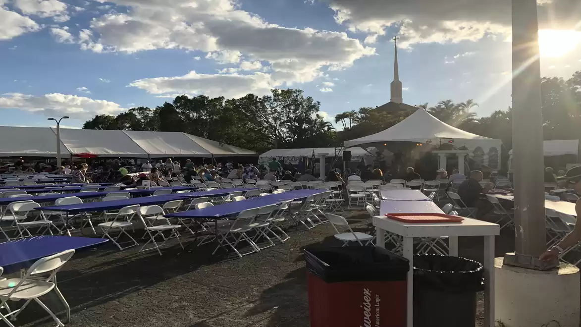 Outdoor event with long tables and chairs under tents. People are gathered around the tables while the sun shines in the sky.